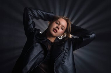 Bir kadın siyah deri ceketle rahatça poz veriyor. Dramatik ışıklandırma dinamik bir arka plan etkisi yaratır. Moda yazıları, giyim markaları veya aksesuar reklamları için..