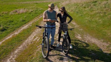 İki bisikletçi, açık mavi gökyüzünün altındaki haritayı kontrol etmek için manzaralı bir kırsal yolda durup güneşli bir maceranın tadını çıkarıyor..