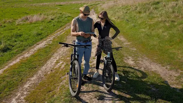 İki bisikletçi, açık mavi gökyüzünün altındaki haritayı kontrol etmek için manzaralı bir kırsal yolda durup güneşli bir maceranın tadını çıkarıyor..