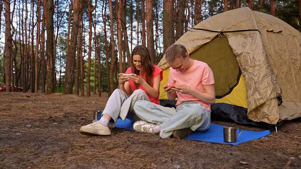 İki arkadaş çadırın yanında oturur, sohbet eder ve akıllı telefonlarıyla oyun oynarlar, ormanda yürüyüş yaparlar..
