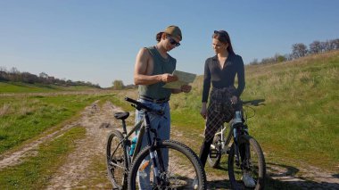 İki bisikletçi, açık mavi gökyüzünün altındaki haritayı kontrol etmek için manzaralı bir kırsal yolda durup güneşli bir maceranın tadını çıkarıyor..