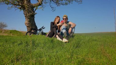 Genç bir çift parkta bir ağacın altında birlikte oturur. Bisikletleriyle güneşli bir günün tadını çıkarıyorlar..