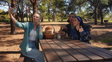 Neşeli bir arkadaş grubu parkta pikniğin tadını çıkarıyor. Uzun ağaçların altında selfie çekiyorlar..