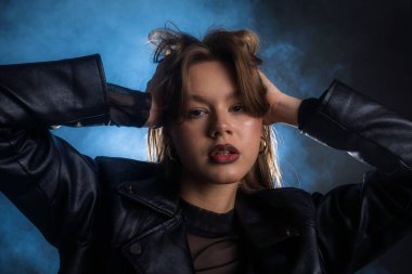 Siyah deri ceket giyen bir kadının çarpıcı portresi, arka plandaki mavi dumana karşı, keskin ve cesur bir moda tarzını vurguluyor. Moda yazıları, giyim markaları veya aksesuar reklamları için..