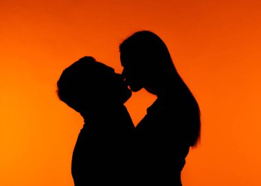 Arka planda canlı turuncu bir gün batımıyla öpüşen bir çiftin silüeti romantik bir anı yakalıyor..