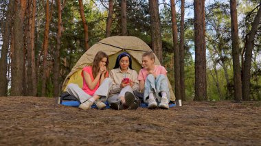 Bir grup arkadaş ormanda kamp yaparken çadırın yanında oturup telefona bakarak endişeli ve endişeli bir şekilde vakit geçiriyorlar..