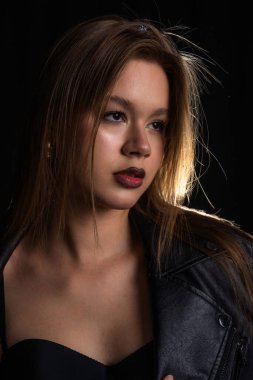 Genç bir kadın, yoğun bir görünüm için yumuşak ve dramatik bir şekilde poz veriyor. Yüzüne ve deri ceketine vurgu yapıyor. Moda yazıları, giyim markaları veya aksesuar reklamları için..