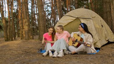 Üç arkadaş ormanda kamp yapmaktan zevk alıyor. Çadırlarının dışında selfie çekiyorlar ve gitar çalıyorlar..