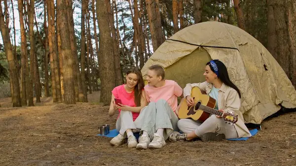 Üç arkadaş ormanda kamp yapmaktan zevk alıyor. Çadırlarının dışında selfie çekiyorlar ve gitar çalıyorlar..