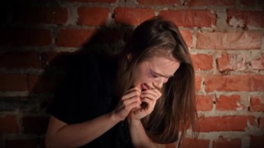 Yaralı genç bir kadın, kasvetli bir ortamda üzüntüsünü ve üzüntüsünü ifade eden bir tuğla duvarın karşısında oturuyor. Yavaş çekim.