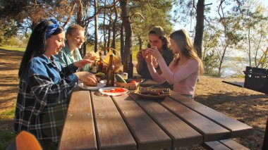 Bir grup arkadaş güneşli bir ormanda pikniğin tadını çıkarıyorlar. Önlerinde yiyecek ve içeceklerle ahşap bir masada oturuyorlar, neşeli ve rahat bir açık hava yaratıyorlar..