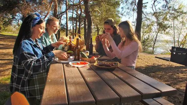 Bir grup arkadaş güneşli bir ormanda pikniğin tadını çıkarıyorlar. Önlerinde yiyecek ve içeceklerle ahşap bir masada oturuyorlar, neşeli ve rahat bir açık hava yaratıyorlar..