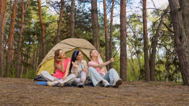 Ormandaki bir çadırın önünde oturan üç arkadaş selfie çubuğuyla anılarını yakalıyor, dışarıda eğleniyorlar..