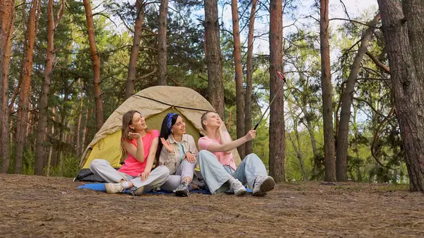 Ormandaki bir çadırın önünde oturan üç arkadaş selfie çubuğuyla anılarını yakalıyor, dışarıda eğleniyorlar..