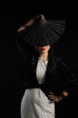 Benekli elbise giymiş sofistike bir kadın siyah bir yelpaze tutuyor, zarafet ve gizem saçıyor..