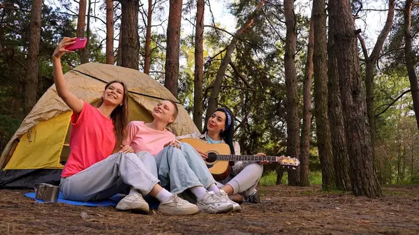 Üç arkadaş ormanda kamp yapmaktan zevk alıyor. Çadırlarının dışında selfie çekiyorlar ve gitar çalıyorlar..