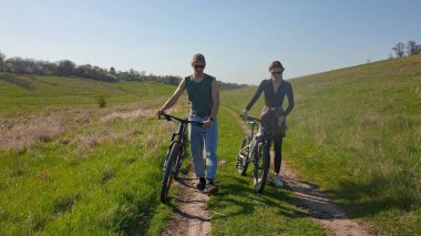 Ağaçlar ve mavi gökyüzüyle çevrili, temiz havanın tadını çıkaran iki kişi açık havada bisiklet sürüyor..