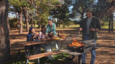 Bir grup arkadaş güneşli bir günün tadını çıkarıyorlar. Uzun ağaçlarla çevrili, pitoresk bir parkta barbekü yapıyorlar..