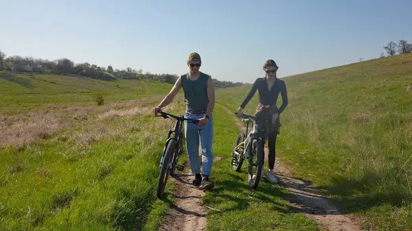 Ağaçlar ve mavi gökyüzüyle çevrili, temiz havanın tadını çıkaran iki kişi açık havada bisiklet sürüyor..