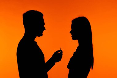 Siluetli çift canlı turuncu bir arka plana karşı duruyor ve romantik bir evlenme teklifi anı yakalıyorlar..