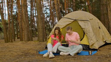 İki arkadaş çadırın yanında oturur, sohbet eder ve akıllı telefonlarıyla oyun oynarlar, ormanda yürüyüş yaparlar..