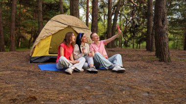 Üç arkadaş ormanda bir çadırın yanında oturmuş, yürüyüş yapıyor ve telefonda görüntülü sohbet yapıyorlar..