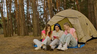 Bir grup arkadaş ormanda kamp yaparken çadırın yanında oturup telefona bakarak endişeli ve endişeli bir şekilde vakit geçiriyorlar..