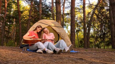 İki arkadaş ormanda bir çadırın yanında dinleniyor gitar çalıyor ve açık havada huzurlu bir ortam seyrediyor..