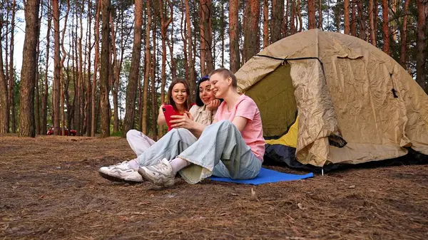 Üç arkadaş ormanda bir çadırın yanında oturmuş, yürüyüş yapıyor ve telefonda görüntülü sohbet yapıyorlar..