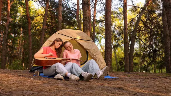 İki arkadaş ormanda bir çadırın yanında dinleniyor gitar çalıyor ve açık havada huzurlu bir ortam seyrediyor..