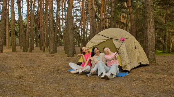 Ormandaki bir çadırın önünde oturan üç arkadaş selfie çubuğuyla anılarını yakalıyor, dışarıda eğleniyorlar..