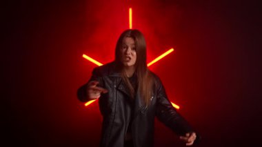 Deri ceketli genç bir kadın cesur, neon ışıklı bir arka plana karşı neşeli bir ifadeyle poz veriyor. Moda yazıları, giyim markaları ya da aksesuar reklamları için. Yavaş çekim.