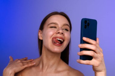 Genç bir kadın mavi akıllı telefonlu bir selfie için şakayla poz veriyor, dilini mavi arka plana doğru uzatıyor..