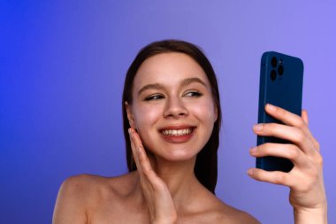 Neşeli genç bir kadın mavi akıllı telefonuyla özçekim yapıyor ve mavi bir arkaplan oluşturuyor..