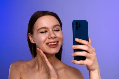 Genç bir kadın, mavi akıllı telefonuyla mor arka planda selfie çekerken gülümsüyor..