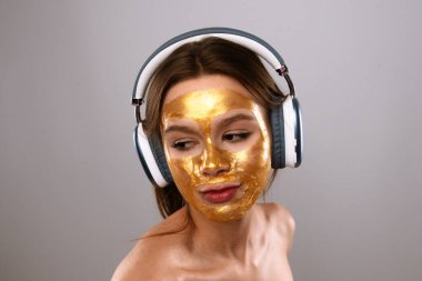 Altın maskeli bir kadın, kulaklıkla müzik dinlerken rahatlıyor ve güzellik tedavisinden zevk alıyor..