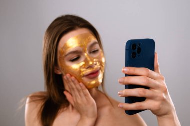 Altın maskeli bir kadın akıllı telefon kullanarak selfie çeker, cilt bakımı ve teknolojiyi ön plana çıkarır..