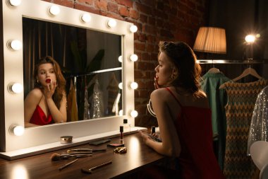 Kırmızı elbiseli bir kadın, şık ışıklandırması ve şık kıyafetleri olan bir soyunma odasına makyaj yapar..