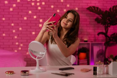 Genç bir kadın, pembe temalı bir odada makyaj malzemeleri ve masanın üzerinde bir aynayla selfie için poz veriyor..