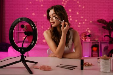 Bir kadın elinde toz fırçasıyla masada oturuyor, sıcak, pembe bir odada kozmetik aletlerle çevrili..