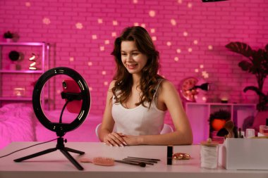 Neşeli bir birey makyaj masasında oturuyor. Güzellik ürünleriyle çevrili. Büyüleyici bir video seansına hazır..