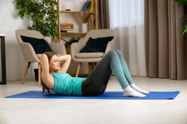 Genç bir kadın, içeride mavi bir minder üzerinde yoga egzersizleri yapıyor, esneklik ve gevşemeye odaklanıyor..