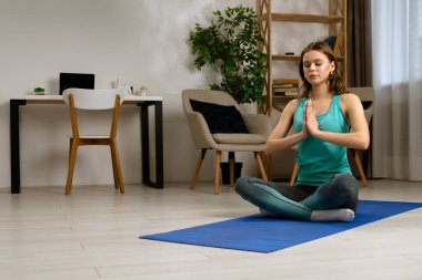 Genç bir kadın, içeride mavi bir minder üzerinde yoga egzersizleri yapıyor, esneklik ve gevşemeye odaklanıyor..