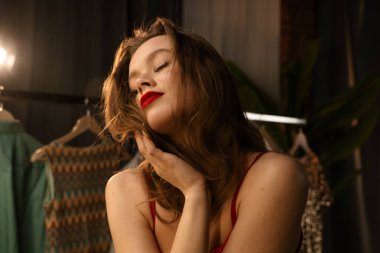 Kırmızı rujlu genç bir kadın, bir askıdaki göz alıcı kıyafetler arasında karar verirken yansımasına hayran kalır..