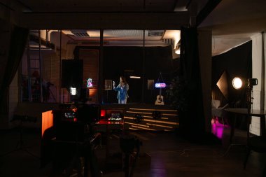 Sahne arkasında. Bir müzisyen karanlık bir stüdyoda canlı performans sergiliyor, neon ışıklarla aydınlatılıyor, ortam ve sanatsal bir atmosfer yaratıyor..
