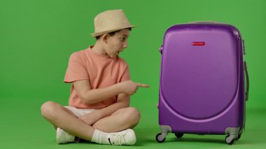 Şapkalı genç bir çocuk mor bavulun yanında oturuyor ve baş parmak işareti yapıyor. Krom anahtar yeşil ekran yeşil ekran stüdyo arka plan modelinde izole edilmiş. Yavaş çekim