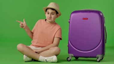 Şapkalı genç çocuk mor bavulun yanında oturuyor ve parmağıyla yeri işaret ediyor, kopyalama alanı. Krom anahtar yeşil ekran yeşil ekran stüdyo arka plan modelinde izole edilmiş. Yavaş çekim