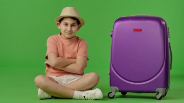 Mor bavulun yanında oturan şapkalı genç çocuk kameraya bakıyor ve gülümsüyor. Krom anahtar yeşil ekran yeşil ekran stüdyo arka plan modelinde izole edilmiş. Yavaş çekim