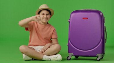 Mor bir bavulla oturan genç bir çocuk yeşil arka planda baş parmak hareketi gösteriyor. Yavaş çekim