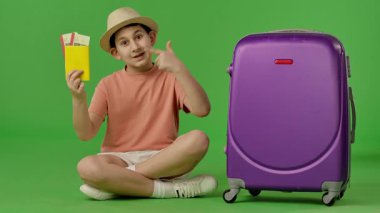 Mor bavulun yanında oturan şapkalı genç çocuk pasaport ve uçak biletleri gösteriyor. Krom anahtar yeşil ekran yeşil ekran stüdyo arka plan modelinde izole edilmiş. Yavaş çekim